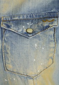 Tascone di giubbotto in denim, blu chiaro con finitura vissuta. Presenta macchie di vernice bianca, un bottone a scatto in metallo e dettagli in cucitura gialla a contrasto.