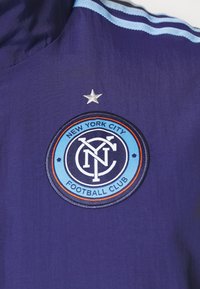 Tissu violet avec un logo circulaire brodé à contour bleu clair, centre noir, accents orange et étoile argentée au-dessus.