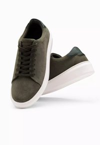Baskets en suède vert foncé avec des semelles en caoutchouc blanc et des lacets plats, présentant un accent en cuir lisse sur le talon et un design minimaliste.