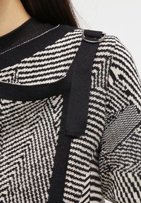 Svartvit mönstrad stickad tröja med ett geometriskt mönster, texturerat tyg och ett svart band med en metallringdetalj på axeln.