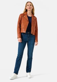 Braune Lederbikerjacke mit silbernen Reißverschlussakzenten, kombiniert mit dunkelblauen Jeans und weißen Sneakers. Modell steht vor einem schlichten Hintergrund.