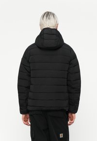 Doudoune noire avec un design matelassé horizontal, dotée d'une capuche, d'une finition texturée et de poignets élastiques. La vue arrière révèle un style minimaliste.