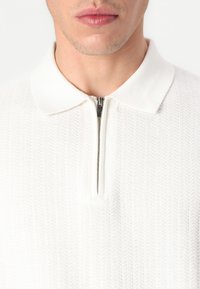 Hvitt ribbestrikket polo-sweatshirt med glidelås i halsen og klassisk krage. Stoffets tekstur er fremtredende; detaljer fremhever håndverket.