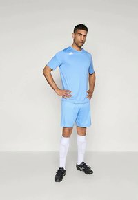 Svetlo modrý atletický dres a šortky, doplnené bielym logom, v kombinácii s čiernymi kopačkami a bielymi vysokými ponožkami. Hladká textúra, priliehavý strih.