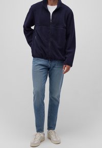 Giacca zip in pile blu navy con superficie texturizzata, tasche frontali e colletto alto, abbinata a jeans azzurri e sneakers crema.