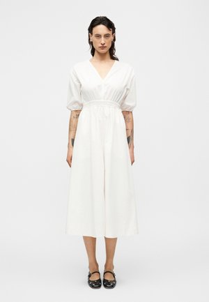 Robe midi en coton blanc avec col en V, manches bouffantes et taille cintrée élastique ; texture lisse et silhouette fluide, associée à des chaussures noires.