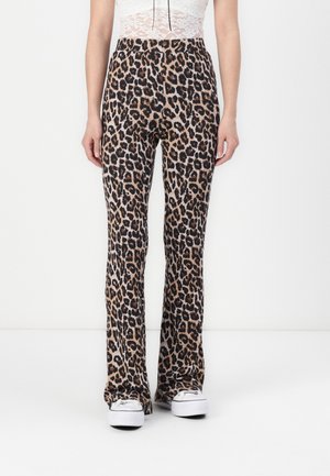 Person står i leopardmønstrede, udsvingede bukser, hvid blondetop og hvide sneakers mod en ensfarvet hvid baggrund.