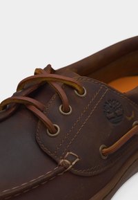 Κοντινό πλάνο καφέ δερμάτινου παπουτσιού τύπου μποτ με δερμάτινα κορδόνια περασμένα μέσα από μεταλλικά κουμπώματα και εγχάρακτο λογότυπο δέντρου στο πλάι.