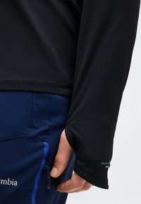 Άτομο που φοράει μαύρο μακρυμάνικο μπλουζάκι με τρύπα για τον αντίχειρα και σκούρο μπλε παντελόνι με ορατή τσέπη με φερμουάρ στο πλάι.