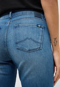 Zili džinsa bikses ar maigu izbalēšanas efektu, tām ir aizmugurējā kabata ar izliekta šuvuma dizainu un zīmola birka pie jostas malas. Gluda tekstūra.