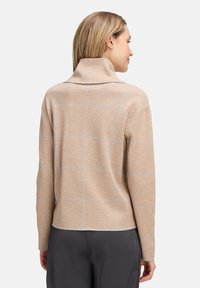 Frau, die von hinten zu sehen ist, trägt einen beigen karierten Pullover mit hohem Kragen und dunkelgraue Hose vor einem einfarbigen Hintergrund.