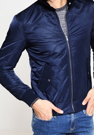 Uomo con una giacca trapuntata blu navy con zip e tasche a bottone, indossa jeans blu e ha le mani nelle tasche.