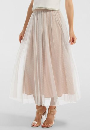 Femme portant une jupe midi beige avec un voile transparent à pois et des sandales à talons transparents sur un fond uni.