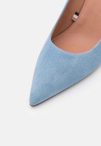 Ljusblå mocka stilett med spetsig tå och slät yta, med en låg skärning och en beige insida. Sulan är svart med minimal slitbana.