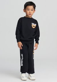 Ragazzo giovane in piedi che indossa una felpa nera e pantaloni con un orsetto e il testo "AWESOME", abbinati a scarpe bianche con velcro.