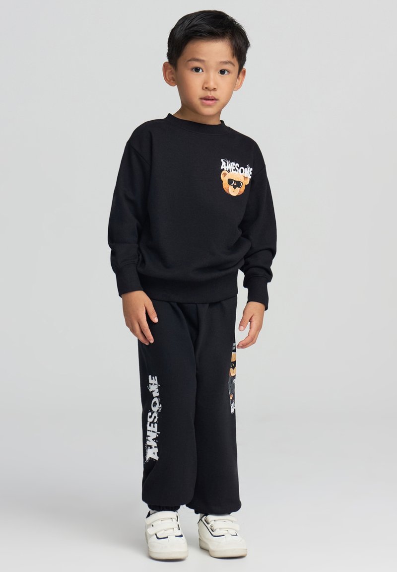 Ragazzo giovane in piedi che indossa una felpa nera e pantaloni con un orsetto e il testo "AWESOME", abbinati a scarpe bianche con velcro.