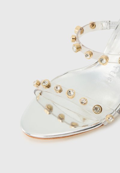 Sandalie cu toc transparent, cu accente metalice aurii și curele transparente, având detalii decorative cu cristale rotunde și strassuri.