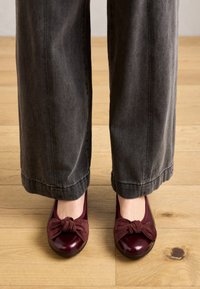Ballerines bordeaux avec un détail en nœud en daim, présentant une finition brillante en vernis et un bout rond, associées à un pantalon large gris.