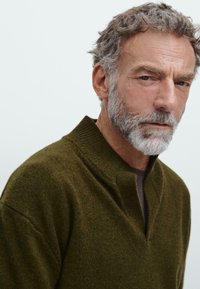 Oude man met krullend grijs haar en een baard, gekleed in een olijfgroen trui met een ingesneden kraag, die iets naar beneden en opzij kijkt.