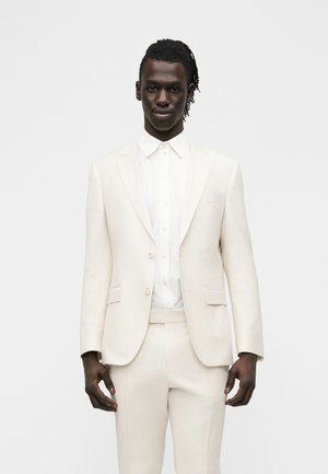 Sötét bőrű, fonott hajú férfi törtfehér öltönyben és fehér gombos ingben, egyszerű világos háttér előtt állva.