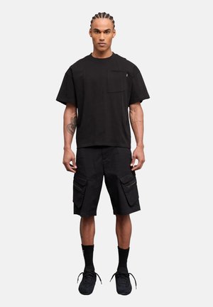 Hombre con trenzas, vestido con una camiseta negra de manga corta y holgada, pantalones cortos cargo negros, calcetines negros y zapatillas negras, de pie frente a un fondo blanco.
