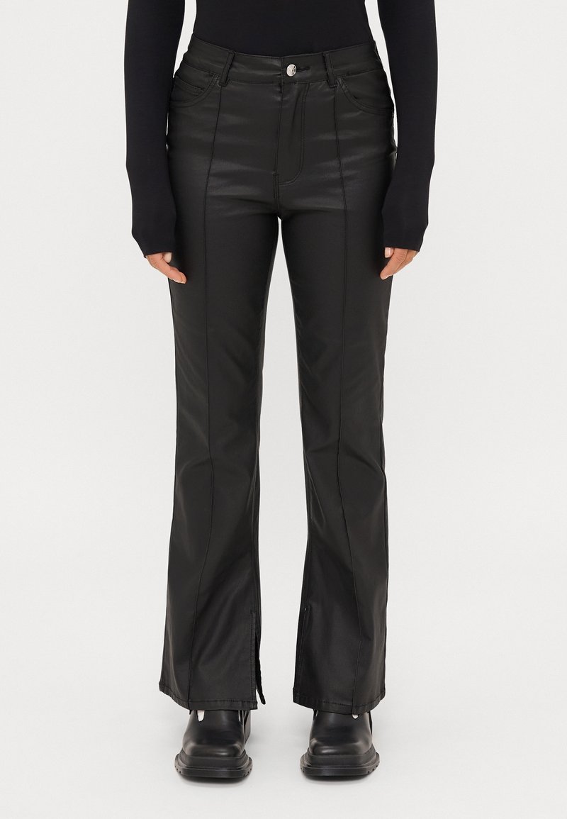 Femme portant un pantalon flare noir taille haute avec coutures à l'avant, fentes latérales aux ourlets, un haut noir à manches longues et des chaussures noires épaisses.