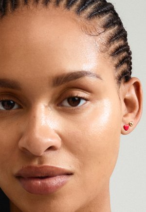 Boucles d'oreilles en or et rouge, avec un petit design en forme de cœur et un accent rond, portées sur l'oreille d'une personne avec des cheveux tressés.
