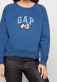 Blå sweatshirt i bomullsblandning med ribbad krage och ärmslut, med en grafik föreställande Mickey Mouse och "GAP" i fet stil.