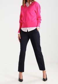 Kobieta stoi z jedną ręką w kieszeni, nosząc jaskraworóżowy sweter z dekoltem w serek, białą koszulę z kołnierzykiem, czarne spodnie i czarne szpilki.