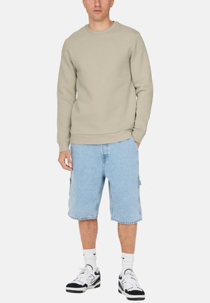 Mann trägt beige langärmliges Sweatshirt, hellblaue knieumspielende Jeansshorts, weiße Nike-Socken und schwarz-weiße Turnschuhe, steht.