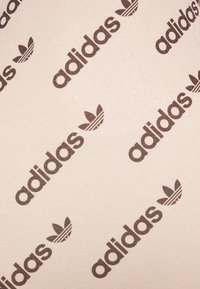 Beige stof met herhaaldelijk donkerbruin "adidas" logo en trefoil afbeelding; gladde textuur met een lichte glans.