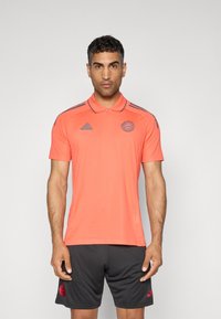 Polo à manches courtes de couleur corail vif avec un col classique et des accents gris, présentant un logo sur la poitrine et des rayures grises sur les épaules.