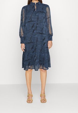 Woman wearing a knee-length navy blue dress with sheer long sleeves, a high ruffled neckline, and abstract swirling patterns.