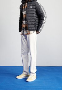 Svart puffjacka med vita ränder, rutig skjorta i brunt och svart, beige byxor samt gula Adidas-sneakers med vita detaljer.