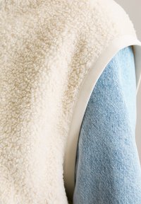 Kremfarget, strukturert shearlingvest med et jevnt, lyseblått denim-underlag. Synlig hvit trim langs armhulen.