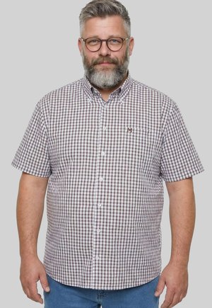 Mann mittleren Alters mit grauem Haar, Bart und Brille, der ein kurzärmeliges kariertes Hemd und blaue Jeans trägt, vor einem hellen Hintergrund stehend.