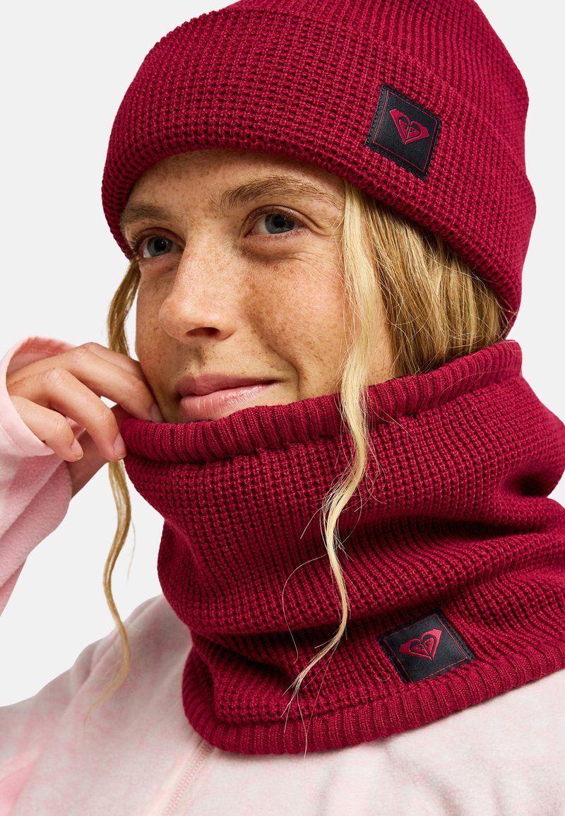Persona joven que lleva un gorro de lana rojo y una braga de cuello a juego, ajustándose la braga con una mano, con piel clara y cabello rubio.
