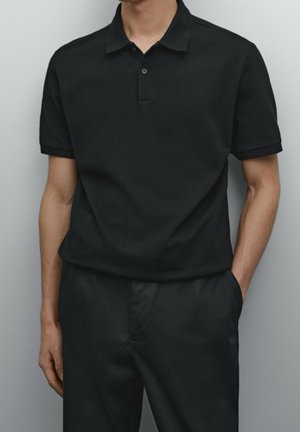 Homme portant un polo noir à manches courtes avec un col boutonné et un pantalon noir, se tenant devant un fond gris clair uni.