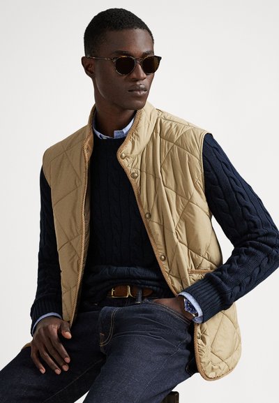 Beżowa pikowana kamizelka, granatowy sweter w warkocze, niebieska koszula na guziki, ciemne dżinsy i okulary przeciwsłoneczne w oprawkach w kolorze żółwia. Klasyczny, warstwowy strój.