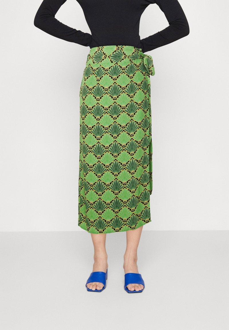 Jupe verte enroulée avec un motif de feuilles répétées, dotée d'un lien à la taille. Portée avec des sandales slides bleues. Tissu en coton avec une texture lisse.