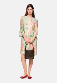 Knee-langes Kleid mit Blumenmuster in Pastellfarben und Glockenärmel; kombiniert mit einer dunkelbraunen Handtasche und roten Schuhen mit Schleifenakzenten.