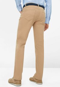 Beige Baumwollhosen mit geradem Bein, zwei Gesäßtaschen und einem strukturierten marineblauen Gürtel. Getragen mit hellbraunen Schuhen.
