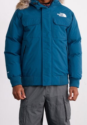 Mann trägt eine blaue isolierte Jacke mit Fellbesatz an der Kapuze und graue Cargohosen, steht vor einem einfarbigen Hintergrund.