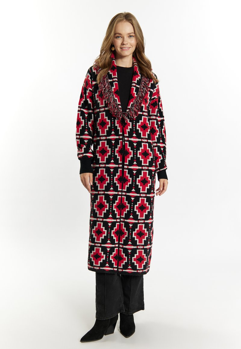 Zwarte en rode gestreepte lange jas met een sjaalkraag, voorzien van franjes. Gedragen over een zwarte top en broek, met zwart met hakken.