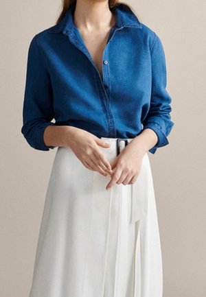 Woman wearing a long-sleeve blue denim shirt tucked into a white pleated skirt with a black belt, hands loosely clasped at waist.