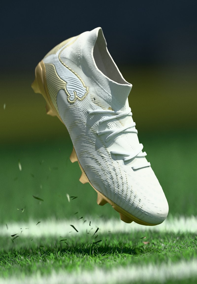 Balti ir aukso spalvos futbolo bateliai, purškiantys žolę žaliame lauke su baltais linijomis, užfiksuoti ore.