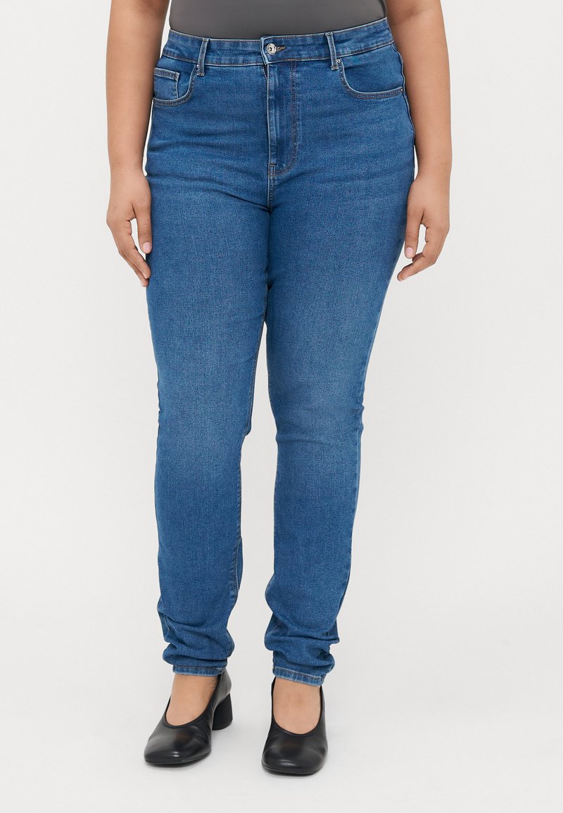 Modre denim hlače z visokim pasom, ozkim krojem in sprednjimi žepi. V kombinaciji s črnimi, okroglimi petami. Gladka tekstura in enakomerna barva.