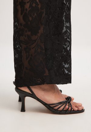 Black open-toe sandals with a slim heel and knotted strap design. The background features intricate floral lace detailing.