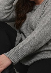 Grå strikket sweater med ribbredder og en rund halsudskæring, der har en blød tekstur. Båret med sorte bukser, der viser en afslappet pasform.