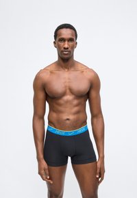 Homme musclé debout devant un fond uni, portant un boxer noir avec une ceinture bleue Nike.